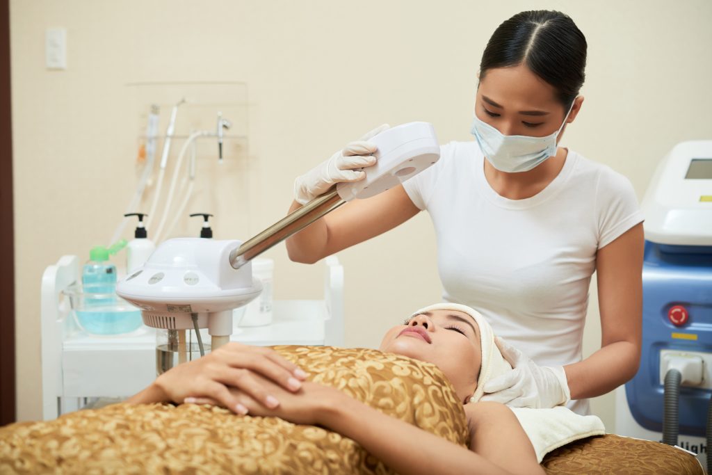 Highly professional Vietnamese cosmetologist wearing rubber gloves and medical face mask standing at treatment table and making necessary preparations for beauty procedure